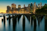 Panoramic cityscape about lower Manhattan's skyscrapers.  The photo taken from brooklyn bridge park. Amazing blue hour view and the sundown on the left side, 
Old pier's columns on the foreground.