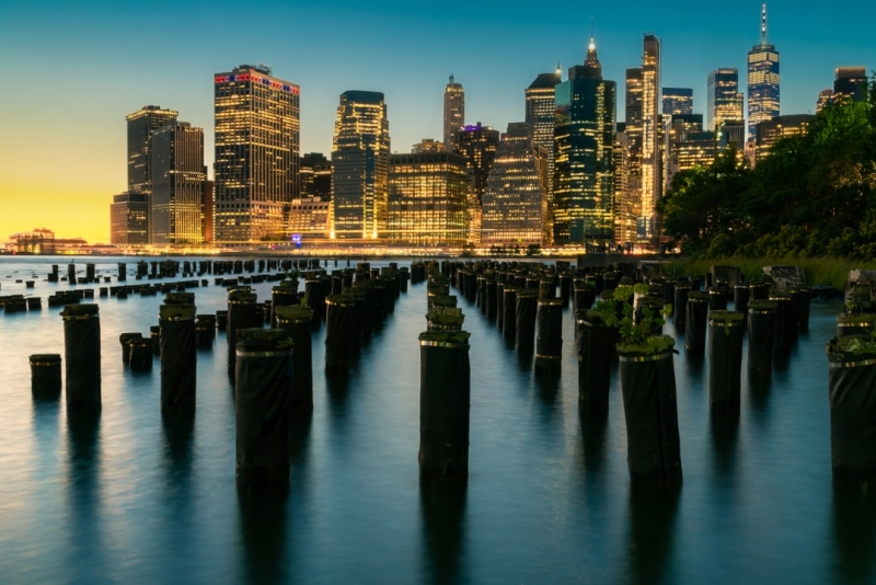 Panoramic cityscape about lower Manhattan's skyscrapers.  The photo taken from brooklyn bridge park. Amazing blue hour view and the sundown on the left side, 
Old pier's columns on the foreground.