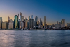 Panoramic cityscape about lower Manhattan's skyscrapers.  The photo taken from brooklyn bridge park. Amazing blue hour view and the sundown on the left side, 
Old pier's columns on the foreground.