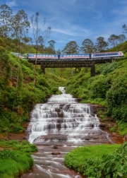A scenic train crosses a metal bridge above the cascading Nanu Oya Waterfalls in Sri Lanka, surrounded by lush greenery and tea plantations, creating a breathtaking view.
