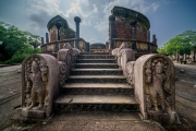 The image depicts the Vatadage, a circular relic house in Polonnaruwa’s Sacred Quadrangle, showcasing intricate stone carvings and ancient architecture under a clear sky