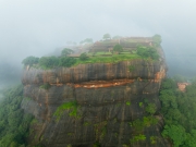 An aerial view of Sigiriya, the ancient rock fortress in Sri Lanka, emerging through the mist, showcasing its lush gardens and historic ruins atop the iconic Lion Rock.
