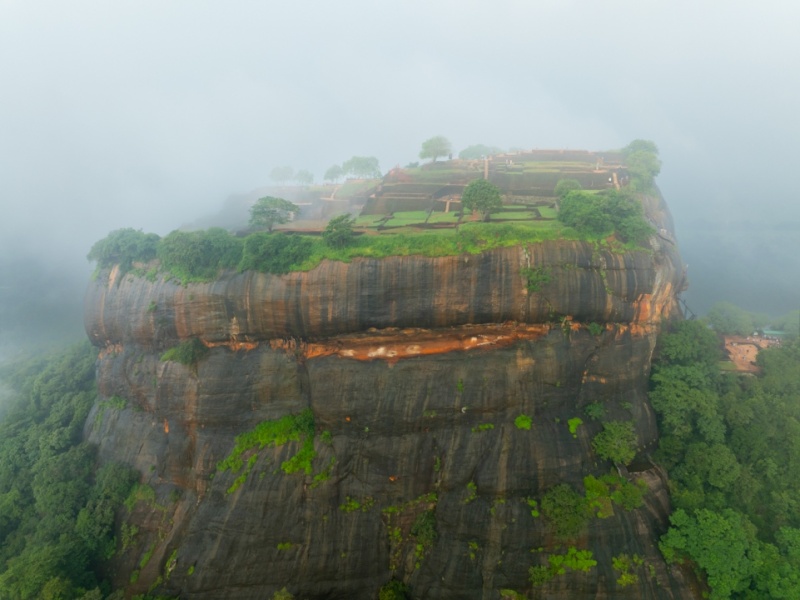An aerial view of Sigiriya, the ancient rock fortress in Sri Lanka, emerging through the mist, showcasing its lush gardens and historic ruins atop the iconic Lion Rock.