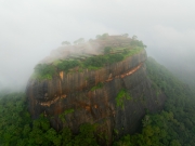 An aerial view of Sigiriya, the ancient rock fortress in Sri Lanka, emerging through the mist, showcasing its lush gardens and historic ruins atop the iconic Lion Rock.