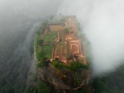 An aerial view of Sigiriya, the ancient rock fortress in Sri Lanka, emerging through the mist, showcasing its lush gardens and historic ruins atop the iconic Lion Rock.