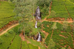 A scenic waterfall cascades through lush tea plantations in Nanu Oya, Sri Lanka, surrounded by rolling green hills and winding dirt paths.