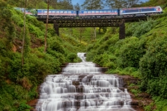 A scenic train crosses a metal bridge above the cascading Nanu Oya Waterfalls in Sri Lanka, surrounded by lush greenery and tea plantations, creating a breathtaking view.